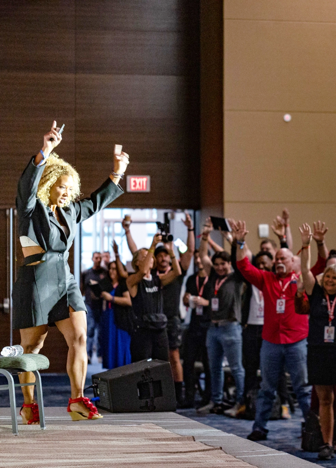 A person on stage in business attire, enthusiastically engaging with an audience who are clapping and raising their hands.