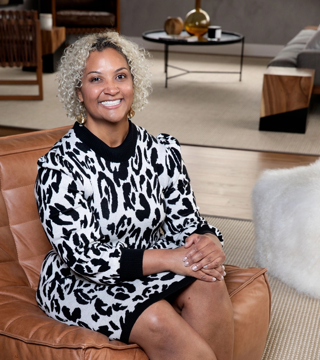 A person with curly hair sits on a leather chair, smiling, wearing a black and white leopard print dress in a modern living room.