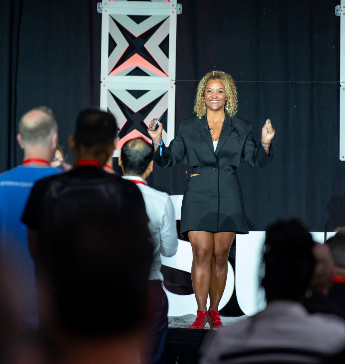 A person in a black outfit speaks on stage to an audience. They hold a microphone and gesture with their hands.