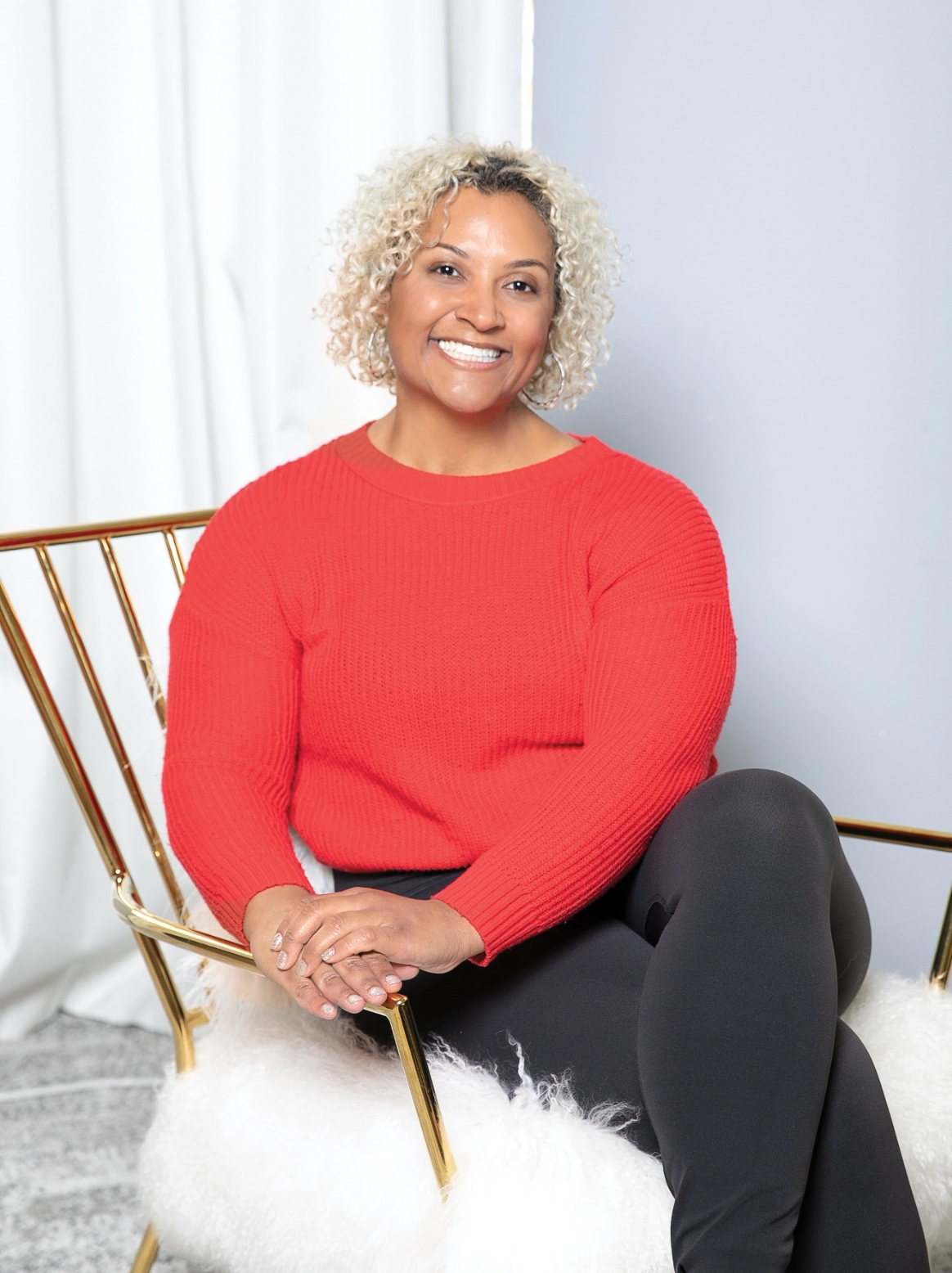 A person with curly hair, wearing a red sweater and black pants, sits on a modern chair, smiling at the camera.