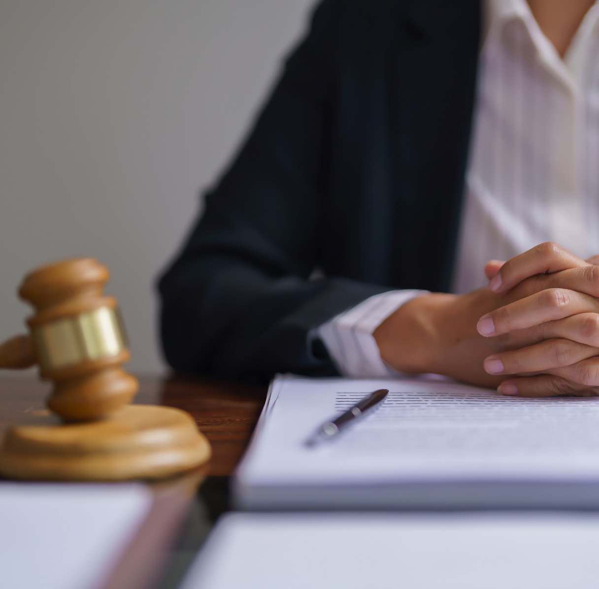 Close-up of a person in a suit sitting at a desk with hands clasped. A judge's gavel and a pen rest on an open document.